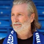 Robbie Savage in a black T-shirt poses with a blue Peterborough scarf