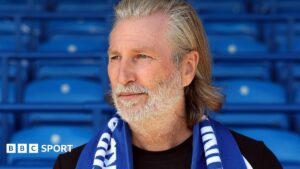 Robbie Savage in a black T-shirt poses with a blue Peterborough scarf