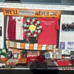A large rectangular glass display case with a black metal frame stands on a grey concrete base outdoors at Manchesterplatz