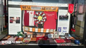 A large rectangular glass display case with a black metal frame stands on a grey concrete base outdoors at Manchesterplatz