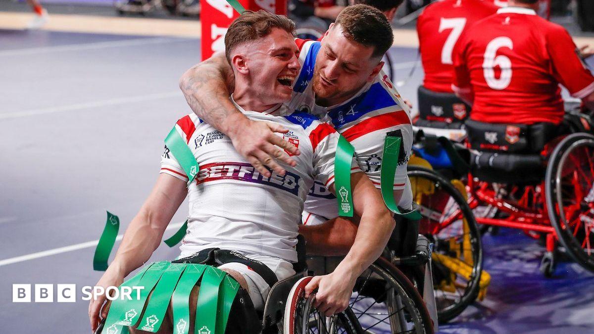 Rob Hawkins, seating in a wheelchair in a white kit with green tags on his shoulders and knees, is hugged from behind by